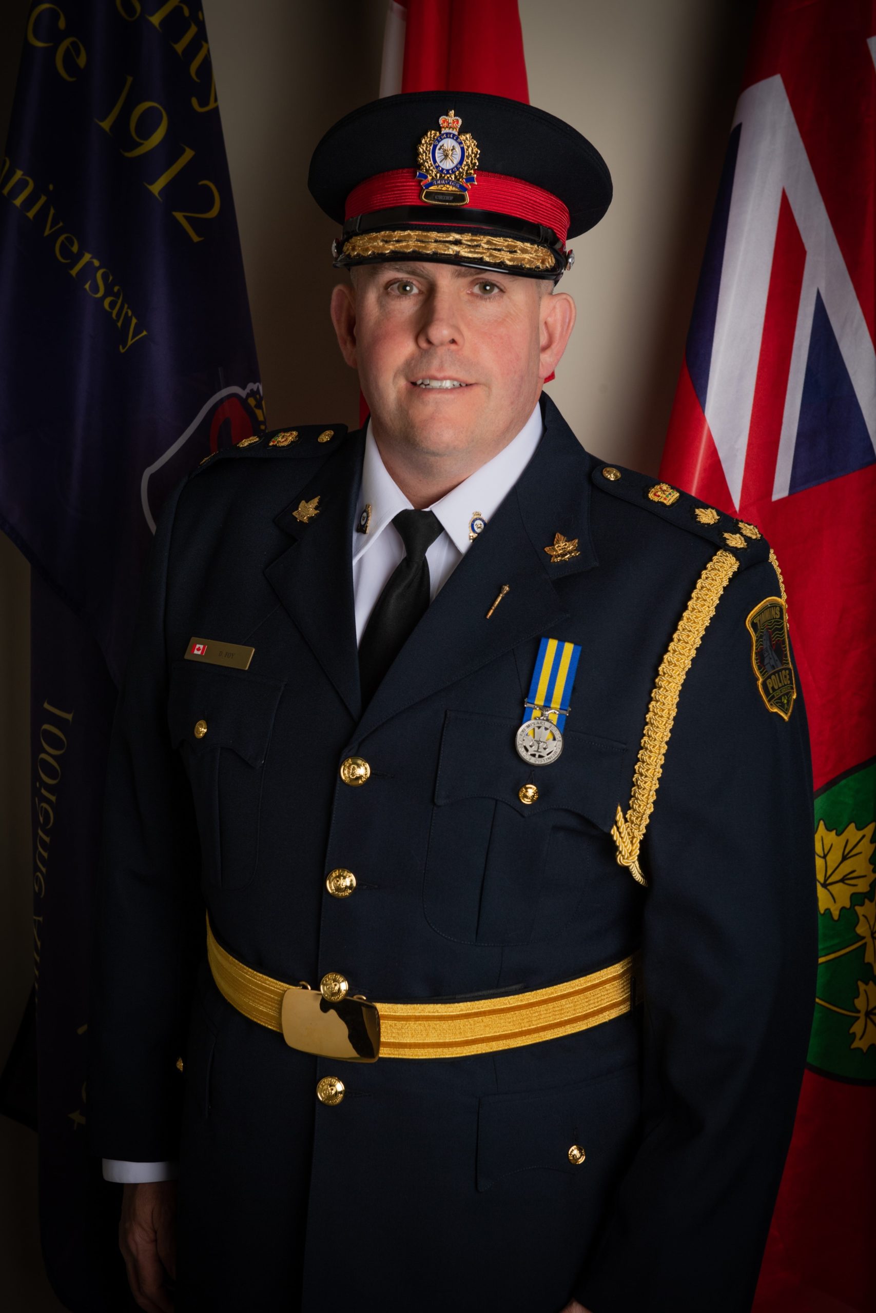 A Timmins Police officer in uniform standing in front of flags.
