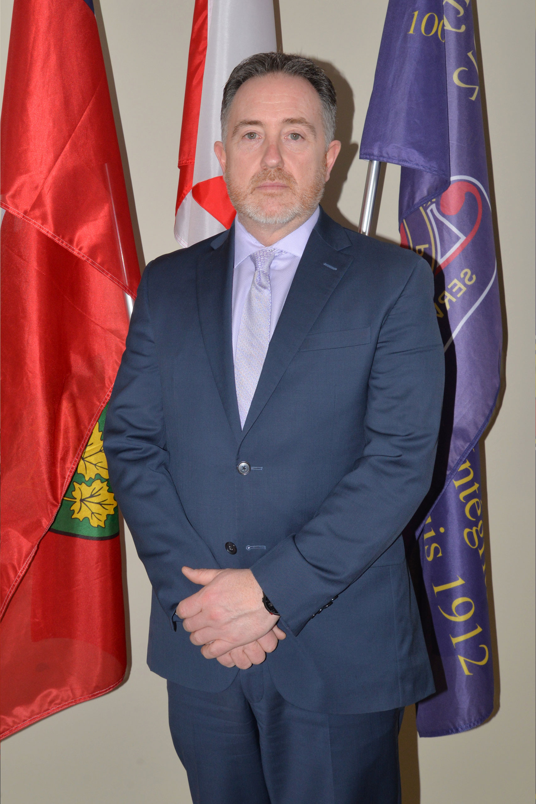 Timmins Police A man in a suit standing in front of Timmins Ontario Police flags.